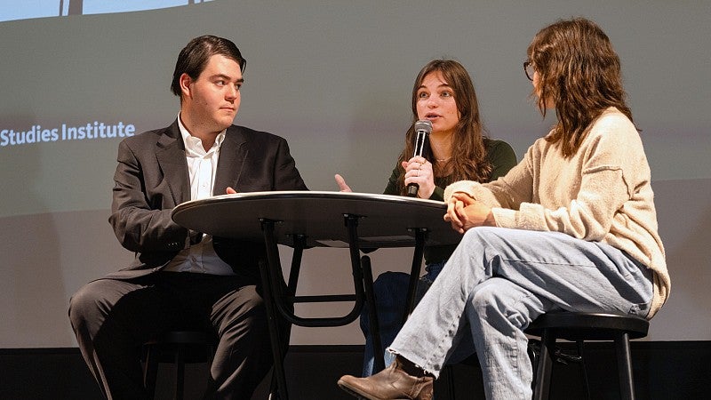 SOJC student Julia Boboc sits at a table on stage and speaks into a microphone while two other students sit on either side of her